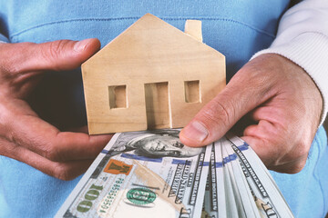 A young man holds a wooden house and a stack of dollars in his hands. The concept of real estate...