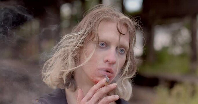 A guy in zombie makeup smokes a cigarette. He puffs out smoke. Bruises under the eyes, bloody mouth. Long narrow hair. Camera movement.