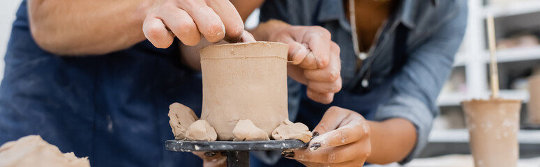 Cropped view of man making clay sculpture near african american craftswoman in pottery workshop, banner.
