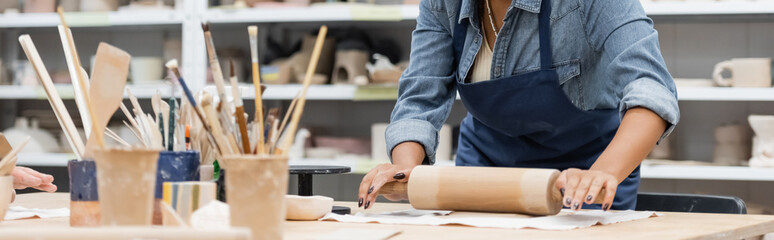 partial view of young african american woman in apron modeling clay with rolling pin, banner.