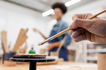 cropped view of man holding shaper in hand near flat clay piece and blurred african american woman.