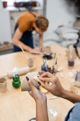 African american woman holding sponge and clay near blurred boyfriend in pottery workshop.