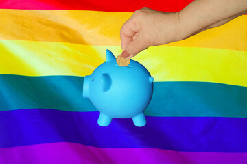 Hand of elderly woman putting coin into piggy bank on LGBT flag background. Hand putting coin to...