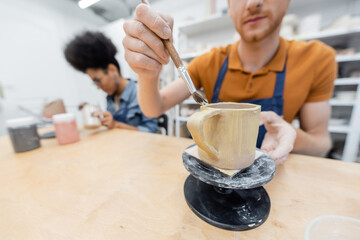 Blurred man painting on ceramic cup near african american girlfriend in pottery studio.