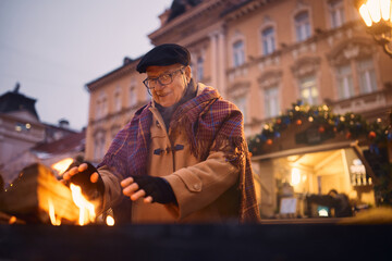 Happy senior man warming up by log fire at Christmas fair during cold winter day.
