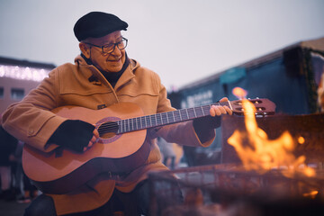 Smiling senior man plays acoustic guitar by bonfire at winter fair.