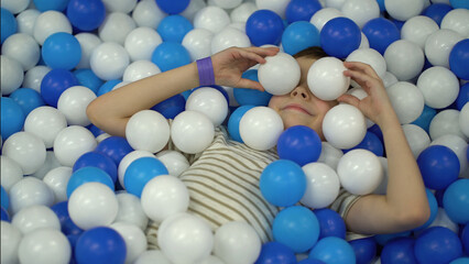 The boy fools around in the pool with colorful plastic balls and throws them - 537849702