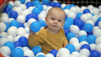 One year old boy sits in a pool with colorful plastic balls and plays with them - 537849761
