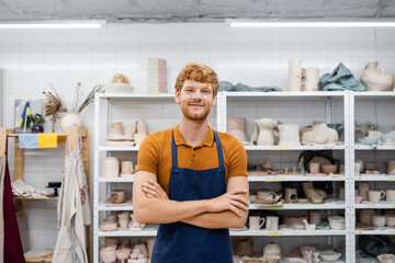 Smiling redhead craftsman looking at camera in pottery workshop.