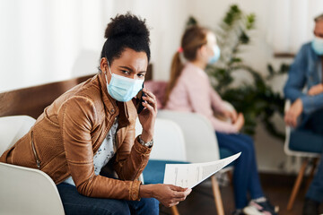 Pensive black woman with face mask talking on cell phone in waiting room at medical clinic.