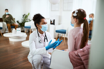 Black doctor with face mask talks to small girl in waiting room at clinic.