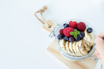 Banana overnight oats with chia seed, blueberries, and raspberries in a jar on white with copy space. Top table view. Plant-based vegan breakfast food concept.