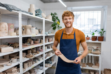positive redhead man in apron holding rectangle shape clay piece in hands near sculptures on rack.