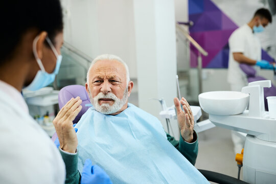 Displeased Senior Man Communicating With Black Female Stomatologist At Dentist's Office.