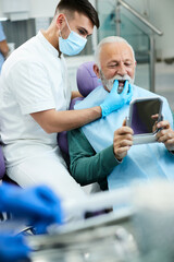 Senior man looking his teeth in mirror while dentist is showing him result of implanting new dentures.