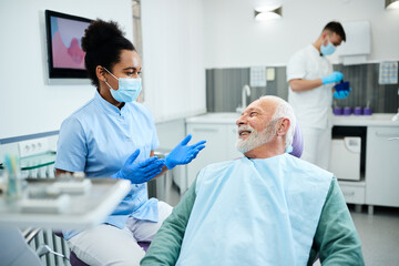 Happy senior man talking to black female dentist during appointment at dental clinic.