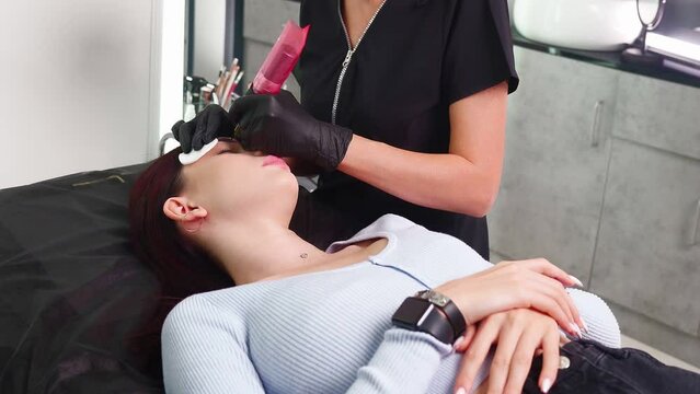 Master makes permanent eyebrow makeup procedure using special needle tattoo machine to woman in beauty salon. Microblading brows tattooing. Dark pigment is injected under skin. Cosmetology procedure