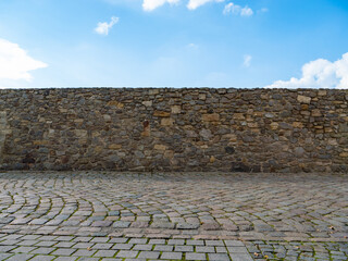 Old stone wall in front of a blue sky with clouds. Empty cobblestone street and an old masonry. Medieval building exterior in a rural area. Abstract background with vintage architecture.