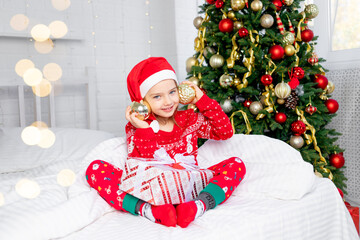 Obraz na płótnie Canvas a child girl with Christmas balls on her ears at the Christmas tree in a red sweater and Santa Claus hat on New Year's Eve in a white bed smiling, fooling around and laughing with a gift