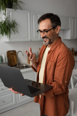 Fototapeta premium happy bearded man in eyeglasses gesturing during video call on laptop in kitchen.