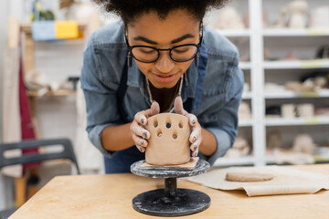 Smiling african american woman in eyeglasses forming clay sculpture in pottery studio.