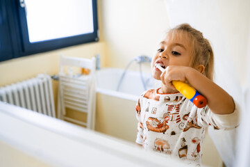 A cute little blonde girl is brushing her teeth with an electric brush, standing in the bathroom, looking in the mirror at her reflection.Close-up, copy the text - obrazy, fototapety, plakaty