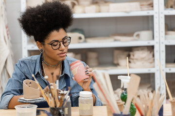 Fototapeta premium African american craftswoman in eyeglasses holding jar with paint near blurred paintbrushes and clay sculpture in pottery studio.