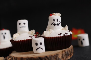 delicious chocolate cupcakes with white cream and marshmallows painted with funny faces on a dark...