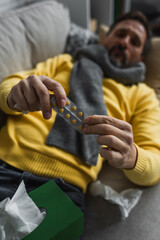diseased man in warm scarf holding pills while lying on couch on blurred background.