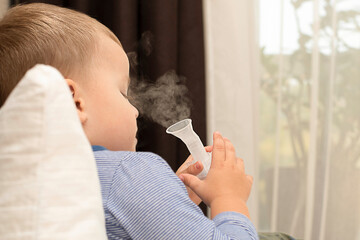 A small caucasian boy lies in bed and breathes cough medicine with an inhaler.