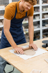 cheerful redhead man in apron modeling clay piece with rolling pin during pottery class.