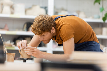 Redhead master in apron creating clay cup in pottery workshop.