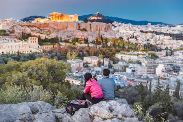 Gardinen Middle aged couple of travellers from Asia sitting at stone admiring fabulous sunset over ancient Athen's city downtown, view at Parthenon and Agora hill, during twilights. Athens, Greece, Europe. © Feel good studio