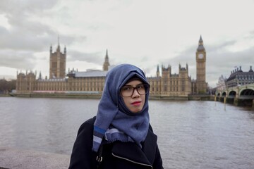Portrait of beautiful young Asian muslim woman wearing hijab and eyeglasses with sad expression on Westminster Bridge with Westminster palace and Big Ben and cloudy sky in background.