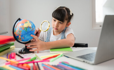 Cute happy Asian child girl student sitting at a desk with a laptop and using a magnifying glass to look and search the map and country on model globe.