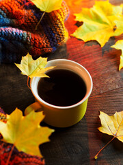 A yellow mug of hot coffee on a wooden table with yellow autumn leaves and a warm knitted scarf