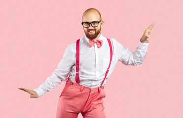 Happy man dancing in studio. Young guy wearing glasses, white shirt, bow tie and pink pants with suspenders dancing isolated on pink background. Handsome Caucasian dancer enjoying music and dancing