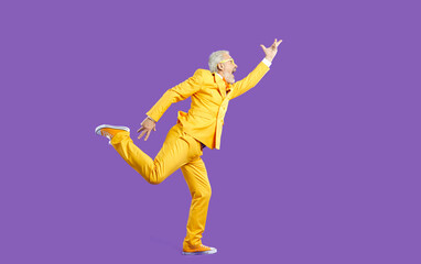 Fototapeta na wymiar Funny senior man dancing, singing and having fun in studio. Side view of happy energetic bearded mature man wearing bright yellow party suit dancing isolated on solid purple colour background