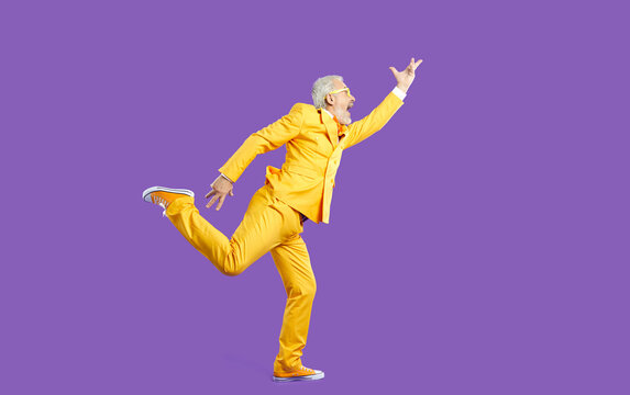 Funny Senior Man Dancing, Singing And Having Fun In Studio. Side View Of Happy Energetic Bearded Mature Man Wearing Bright Yellow Party Suit Dancing Isolated On Solid Purple Colour Background