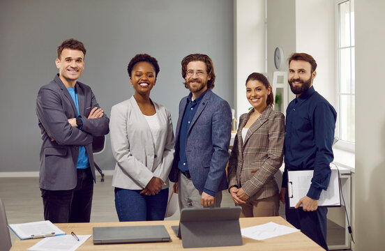 Multiethnic People On Board Of Directors Of Corporation Stand With Smile Next To Table With Documents Posing For Website About Company Or Startup Located In Modern Office. Business Success