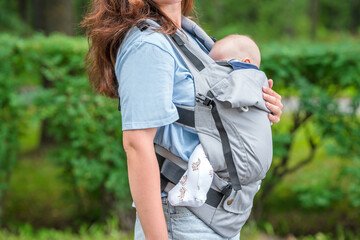 A young mother walks with a baby sleeping in a sling backpack in a green summer park