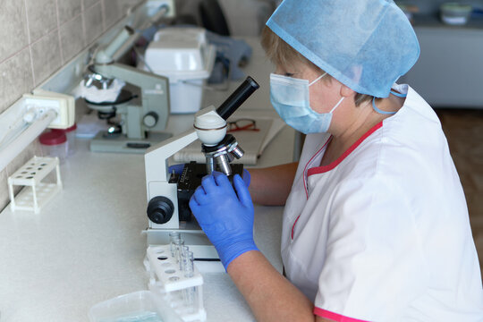 Woman Scientist Wearing Face Mask And Protective Gloves Working In Laboratory Looking Through Microscope. Female Researcher Doing Investigations Using Special Blood Test Equipment With Test Tubes