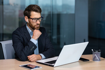 Fototapeta premium Serious thinking mature businessman working inside modern office building, senior boss in business suit and glasses working sitting at laptop, man with beard thinking about future strategy plan.