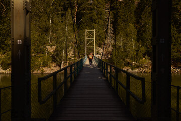 Man with backpack walking across a narrow suspension bridge over a river in a lush forest during golden hour. Cinematic view of outdoor adventure, hiking, and exploration in nature.