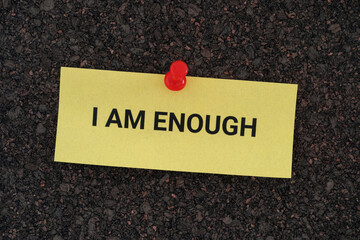 A yellow paper note with the phrase I AM ENOUGH on it pinned to a cork board