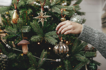 Hands decorating christmas tree with stylish bauble in atmospheric festive room. Merry Christmas! Winter holidays preparation. Woman in cozy sweater putting vintage toy on tree
