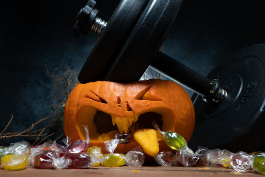 Dumbbell barbell weight plates crushing carved Halloween pumpkin Jack-o'-lantern head. Candy spills out of the mouth. Healthy diet, fitness lifestyle composition. Gym workout sport training concept.