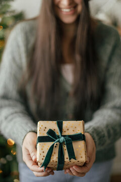 Stylish christmas gift in hands against christmas tree with lights. Merry Christmas and Happy Holidays! Woman in cozy sweater holding wrapped present close up in atmospheric festive room