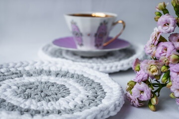a purple porcelain coffee mug on a pink plate on a crocheted coaster and pink flowers in the foreground. Cozy kitchen background. Glamorous lifestyle poster. Place for text