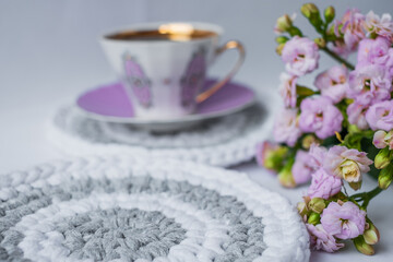 Obraz na płótnie Canvas a purple porcelain coffee mug on a pink plate on a crocheted coaster and pink flowers in the foreground. Cozy kitchen background. Glamorous lifestyle poster. Place for text